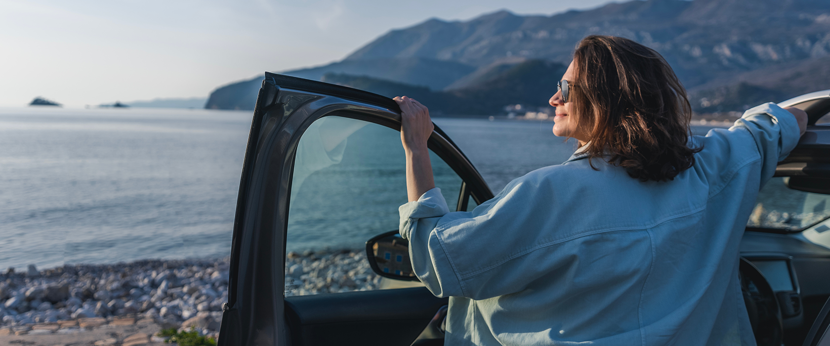 Eine Frau mit Sonnenbrille steht mit dem Rücken zur Kamera an einer offenen Autotür und guckt auf ein Gewässer
